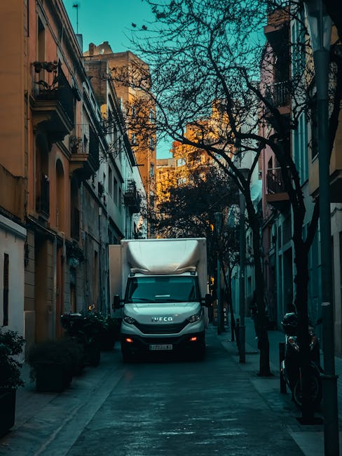 A white IVECO box truck is parked on a narrow urban street with tall, closely spaced buildings on either side, featuring residential balconies and ornate architecture. The truck is positioned facing directly toward the camera, with its front end occupying most of the scene. The street is lined with leafless trees, and the pavement shows a series of stacked moving boxes wrapped in brown packing paper and plastic, along with some furniture covered in protective blankets, indicating a home relocation process. Visible around the scene are small potted plants, motorbikes parked along the sidewalk, and various street fixtures such as bollards and overhead wires. The lighting suggests late afternoon or early evening, creating a warm glow on the buildings in the background. This setting captures the loading of household items into the vehicle by Man With a Van Kew, highlighting a professional furniture transport and packing process typical in house removals and professional relocation services.
