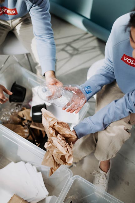 Two individuals are engaged in a home relocation process, handling packing materials inside a property. One person is wearing a light blue long-sleeved shirt and beige trousers, holding a plastic water bottle with a blue cap and wrapping it with bubbles for protection, while the other is wearing a light grey hoodie and beige trousers, placing items into a plastic storage container. The container is partially filled with crumpled brown paper and other packing supplies, positioned on the floor near a stack of boxes and packing paper. The scene takes place on a wooden or laminate floor, with a modern sitting room visible in the background, including a grey sofa. This activity is part of a furniture transport or packing and moving stage often associated with professional removal services, such as those offered by Man With a Van Kew, supporting efficient house removals and recycling routes.