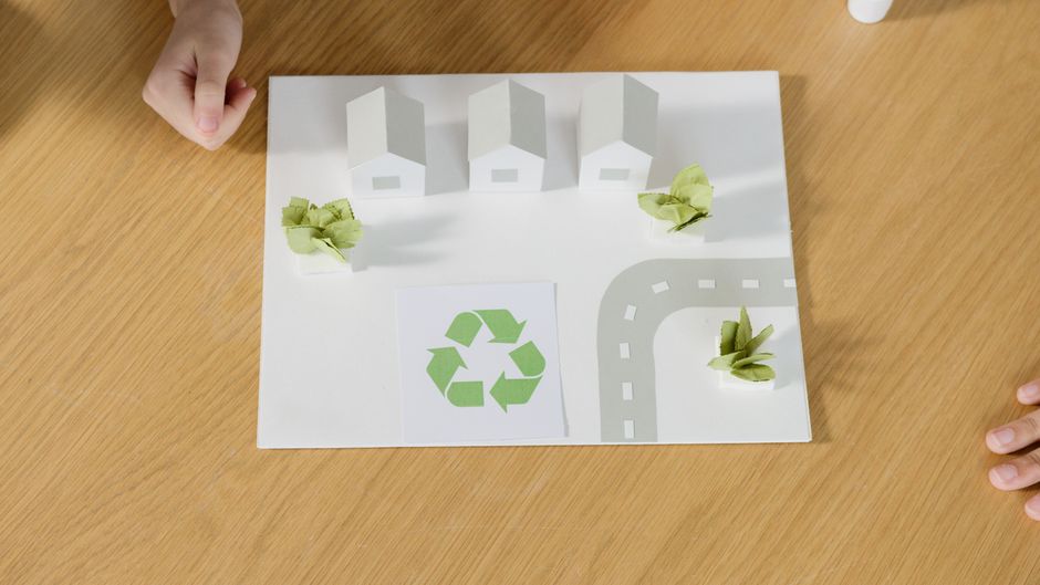 A top-down view of a house relocation planning model on a wooden table, featuring three white paper houses positioned at the top, representing residential units, with small green paper trees and bushes placed around them to depict landscaping. Below the houses, there is a printed green recycling symbol on a white paper square, indicating sustainable disposal or donation efforts during the move. To the right of the recycling symbol, a curved grey paper road with white dashed lines is illustrated, simulating a street layout. Two small green paper trees are positioned along the road, enhancing the layout's realism. A hand is visible at the top left corner, and another at the bottom right corner, suggesting active involvement in the moving planning process. The scene is set on a clean, light-colored wooden surface, with the model used to illustrate a house relocation and eco-friendly disposal process, as part of a home removal project managed by Man With a Van Kew.