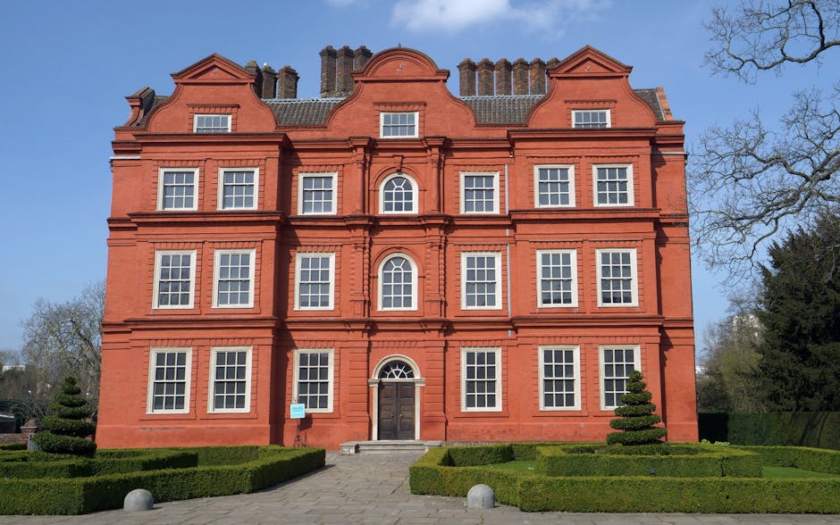 A three-story red-brick building with decorative architectural details and multiple evenly spaced white-framed windows, located in a landscaped area with neatly trimmed hedges and small spiraled topiary bushes. The entrance features a dark wooden door accessed by a small set of stone steps. The building is situated outdoors under a clear blue sky, with a tall tree on the right and evenly spaced chimneys on the roof. The scene suggests a well-maintained residential or institutional property, possibly related to house removals or property relocation services, with external parts visible that could be relevant for furniture transport and home moving logistics.