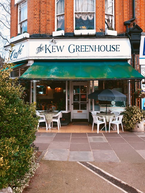 The image shows the exterior of a brick building housing 'The Kew Greenhouse' café, with a white signboard displaying the establishment's name in blue and black lettering. In front of the café, there is a small outdoor seating area on a paved pavement, featuring several white plastic chairs and matching tables, one of which is shaded by a large black umbrella. To the left of the seating area, a bush with green foliage partially obscures the view. The entrance to the café is beneath a green awning, leading inside where interior lighting is visible through the windows. The scene is set during daylight hours, and the overall atmosphere is calm and inviting, typical of a location involved in home relocation or furniture transport logistics when coordinating loading and unloading during a move. The image captures an exterior scene relevant to professional removals and moving services, as provided by Man With a Van Kew, emphasizing an accessible, well-maintained commercial property exterior. 