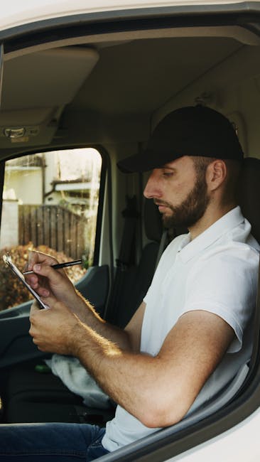 A man with a dark beard and wearing a black cap and white T-shirt is seated inside a moving van, looking at a smartphone held in his right hand. His left arm rests on the vehicle's door frame, which is open, revealing the interior of the van. The background through the window shows a garden with a fence and some plants, indicating he is preparing for or coordinating a home relocation. The van's interior ceiling is visible, along with the window frame and part of the vehicle's headliner. [COMPANY_NAME], Man With a Van Kew, provides professional removals and furniture transport services, supporting efficient packing and loading processes for house and office moves, as reflected by this loading or planning scene.
