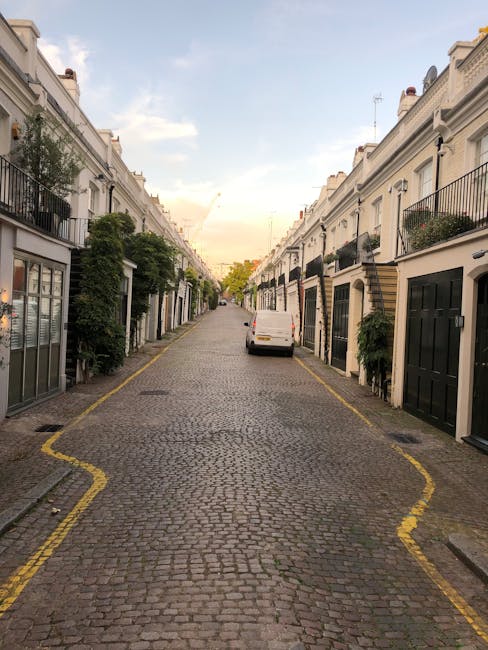 A narrow residential street with cobbled pavement lined on both sides by white terraced houses featuring black and brown doors, some with small front gardens and potted plants. A white van is parked on the right side of the street near the curb, with the rear doors slightly open, suggesting loading or unloading in progress. The street is marked with yellow lines along the edges, indicating parking restrictions. The sky above is partly cloudy with soft evening light illuminating the scene. This setting exemplifies a typical environment where house removals and furniture transport may require careful navigation due to limited vehicle access, as discussed in Man With a Van Kew's guide on narrow street vehicle access for Kew removals estates.