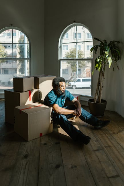 A man with dark curly hair and wearing a blue T-shirt and dark jeans is sitting on a wooden floor inside a room with large, arched windows that allow natural daylight to illuminate the space. The room contains several packed cardboard boxes, some stacked on top of each other, sealed with red packing tape, indicating recent or ongoing packing for a home relocation. A tall potted plant with broad green leaves is positioned in the corner near the windows. Outside the windows, a street scene with parked cars and residential buildings is visible, suggesting an urban environment. The image captures the quiet moment during packing and moving preparations, which relate to furniture transport and packing processes involved in house removals, as represented by the setting at [DOMAIN]. This scene reflects the logistical aspects of professional removals services provided by [COMPANY_NAME], emphasizing the unpacking, packing, and loading phases typical in an effective house move.