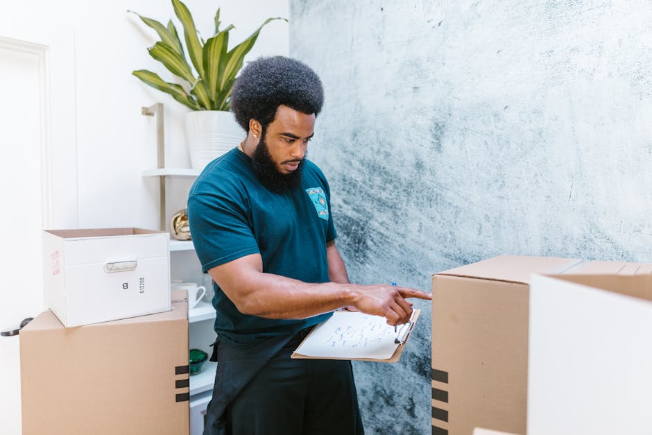 A man with a dark skin tone and an afro hairstyle, dressed in a navy blue T-shirt and dark trousers, is inside a spacious, well-lit room with white walls and wooden flooring, during a home relocation or furniture transport process. He is holding a cardboard box, sealed with red tape, in his right hand, while standing next to two other boxes placed on the floor—one directly beneath his hand, and the other slightly in front of him. The room features a large arched window behind him, allowing natural light to illuminate the scene, with a view of the outside and partly visible window frames. The man appears to be preparing or in the midst of packing or unpacking, consistent with furniture and packing materials involved in house removals. He is wearing a work apron tied around his waist, with additional boxes and packing supplies possibly supporting a professional move, which aligns with services provided by Man With a Van Kew. The environment suggests an organized and careful packing or loading process during a home move, with attention to packaging materials and furniture transport.