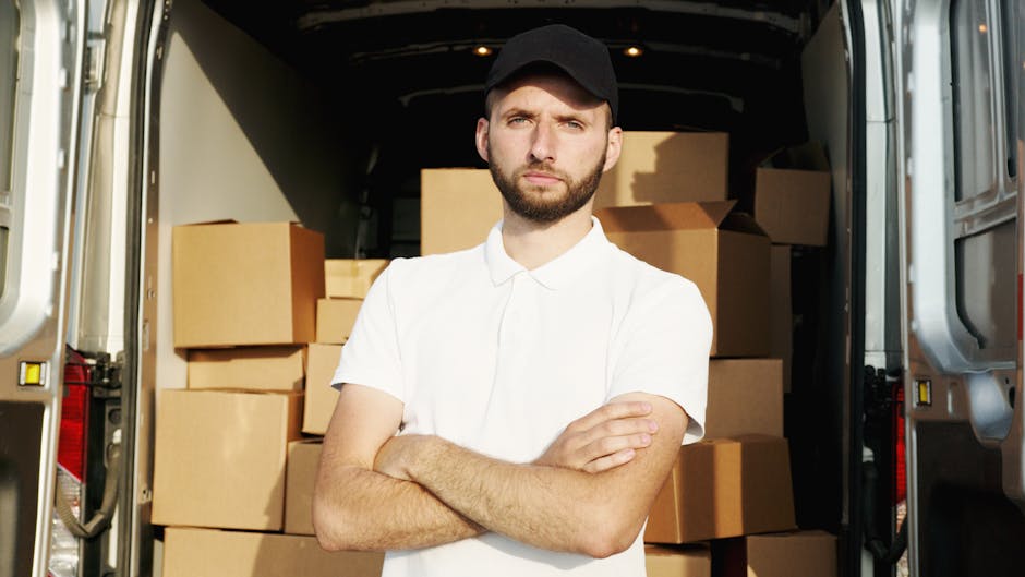 A man with a serious expression and a beard, wearing a black cap and a white polo shirt, stands with his arms crossed inside the open rear of a large box truck. Behind him, the truck's interior is visible, filled with neatly stacked cardboard moving boxes of various sizes, some wrapped in plastic or packing material. The truck is parked on a street, with a pavement visible outside. The scene reflects a home relocation or furniture transport process, typical of professional removals services such as those offered by Man With a Van Kew, involving loading or unloading boxes during a same day house move in Kew TW9 area.