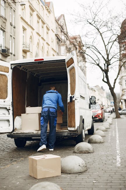 A man wearing a blue jacket and dark trousers is seen loading large cardboard boxes and furniture into the back of a white box truck parked on a city street. The truck's rear doors are open, revealing an empty interior with wooden paneling. The scene takes place on a cobblestone pavement lined with large, rounded concrete bollards and parked cars, with historic multi-storey buildings and leafless trees visible in the background. The background also shows a partly cloudy sky, and the area is illuminated with natural daylight. The activity appears to be part of a home relocation or furniture transport process, with the man handling packaging materials and preparing items for moving, aligning with professional removals and packing services offered by companies like Man With a Van Kew.