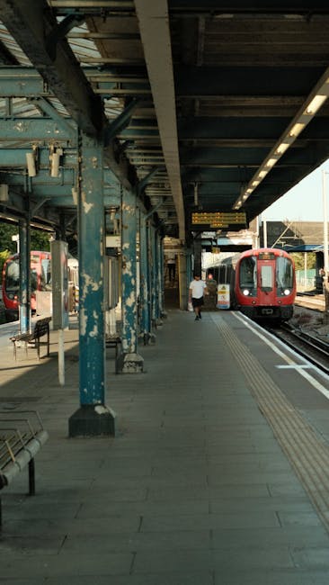 A train station platform with weathered blue painted metal columns supporting an overhead canopy made of wooden beams and dark roofing material. The platform features tactile paving strips along the edge for passenger safety. A red train with a sleek modern design is stopped on the right side of the image, with its doors open for boarding. A single passenger dressed in casual clothing is walking along the platform towards the train. There are benches and other station features visible along the left side, and the station environment appears well-lit by natural light. This scene depicts a typical urban transportation hub suitable for home relocation and moving services such as those offered by Man With a Van Kew, especially relating to packing, loading, and transporting belongings via train as part of a comprehensive house or flat removal process near Kew Gardens station.