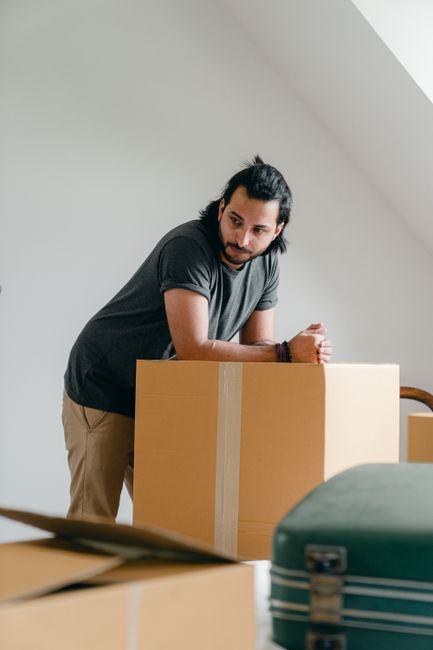 A man with dark hair tied back, wearing a dark grey t-shirt and light beige trousers, is standing indoors beside a large cardboard box wrapped with clear packing tape, preparing for a home relocation. The room features a sloped white ceiling with natural light illuminating the scene. In the foreground, there are additional packed boxes and a green suitcase, indicative of packing and moving procedures. The man appears to be checking or securing the box as part of a furniture transport or house removals process. This image, associated with Man With a Van Kew, illustrates the packing and loading phase typical of professional moving services, emphasizing careful handling of boxed belongings before transport to a new property.