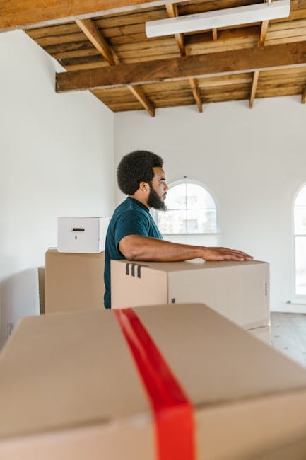 A man with an afro hairstyle and beard, wearing a dark teal t-shirt, is standing inside a room with a high, exposed wooden beam ceiling and white walls. He is positioned behind a stack of cardboard moving boxes, some sealed with red packing tape, while he appears to be preparing for a home relocation. The boxes are of various sizes, and some are placed on top of each other near a window with white frames, allowing natural light to illuminate the space. The room’s minimal decor and natural lighting suggest an early stage of packing and moving preparations. The scene is part of a furniture transport or packing and moving process, as typical in professional removals, with the focus on handling and organizing household items ahead of an upcoming house move, as seen in services offered by companies like Man With a Van Kew.